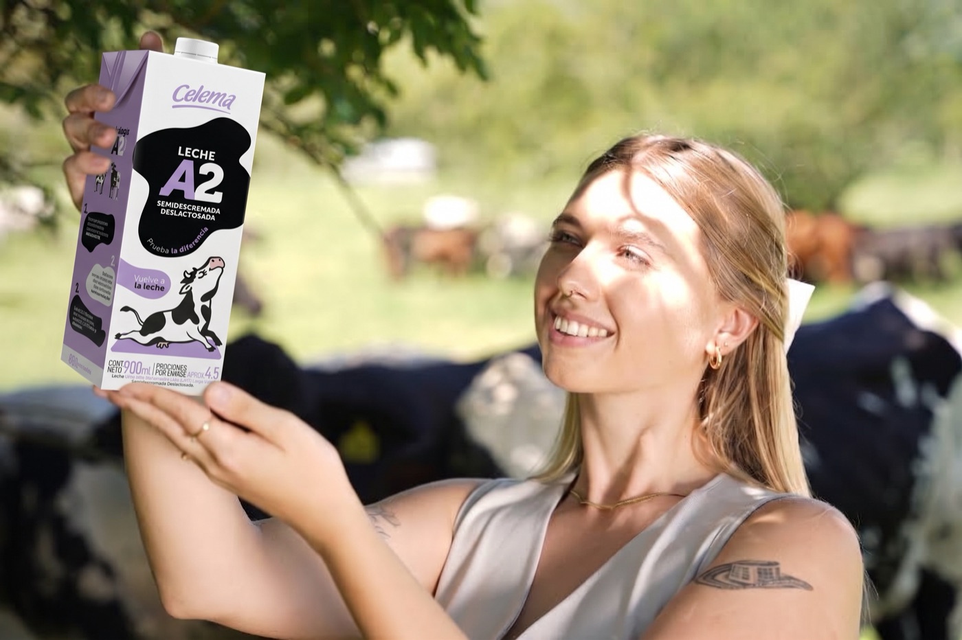 Ganadera sonriente sosteniendo en alto un empaque de Leche Celema A2, con vacas al fondo